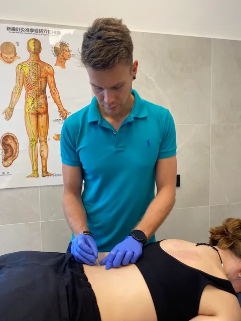 Paweł Grajewski wprowadza igłę akupunkturową na tle planszy medycyny chińskiej